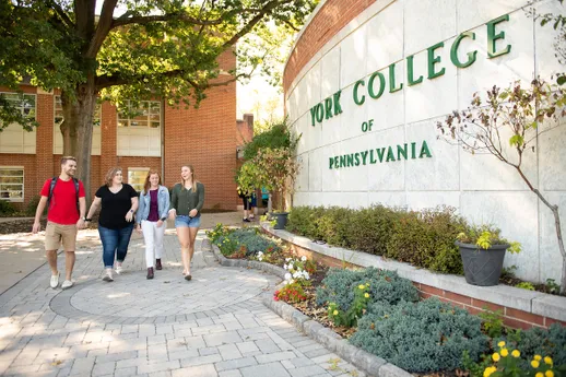 Students walk on campus past a large, scenic wall with an embedded York College of Pennsylvania logo