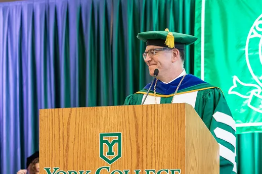 President Thomas Burns smiles while glancing off-stage as he stands at the podium in full academic regalia during Convocation.