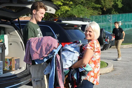A mother helps her late-teenaged son carry a box of clothes out of an SUV.
