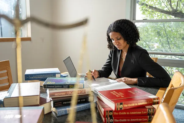 A student in a blazer sits at a desk, surrounded by books and her laptop as she studies. The edge of the scales of justice are visible in a blurred foreground.