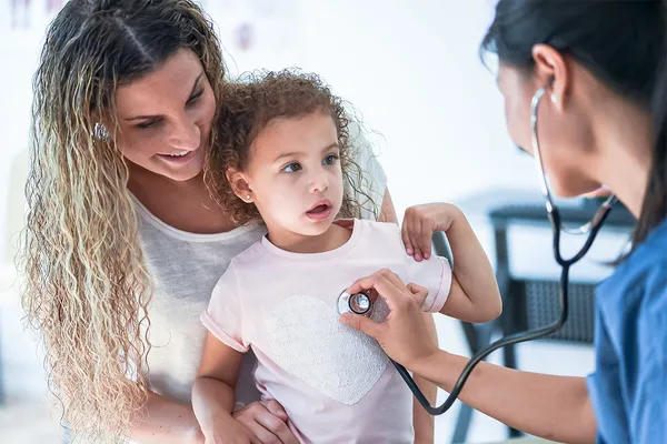 A nurse practitioner in scrubs uses a stethoscope to listen to the heartbeat of a child sitting on their mother's lap.