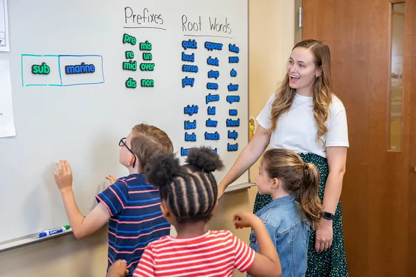 A teacher stands by a whiteboard in a classroom and watches as three students work with magnets featuring prefixes and root words.