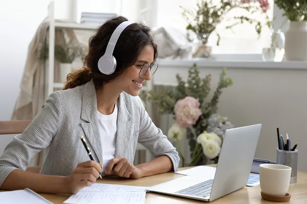 A woman in a blazer wears headphones and takes notes as she participates in an online class with a laptop.