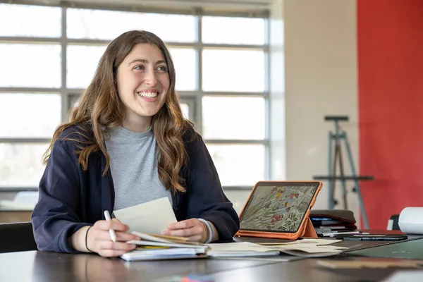 A student in the graphic design studio smiles at something off-camera while sitting at a table covered with papers and a tablet displaying digital art, and a series of pens and pencils.