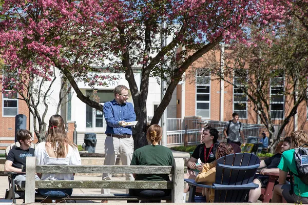 A professor stands in the campus quad, under a blooming purple tree on a sunny day. Students sit in Adirondack chairs and look on during this outdoor class session.