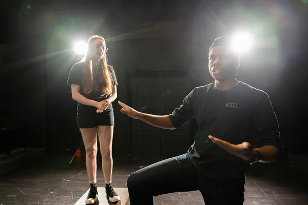 Two students act out a scene in the black box theatre, stage lights illuminate them from behind.