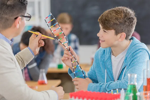 A high school student holds a plastic model of a DNA helix as his teacher looks on, pointing at areas of the model with his pencil.