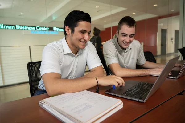 Two students sit side by side in a classroom, with a notebook and laptop in front of them and a NASDAQ ticker visible in the background.
