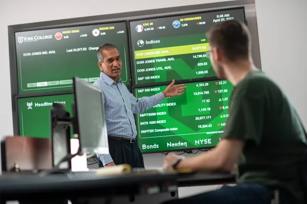 A professor points at a large group of four screens showing data and stock information as a student looks on from his seat at a computer desk