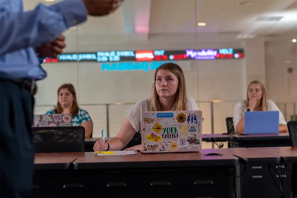 Three students take notes while listening to a professor lecture at the front of the class. Background shows a NASDAQ ticker screen on the wall.