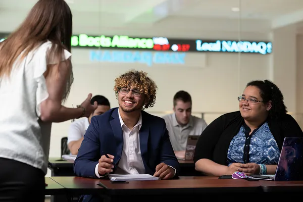 Two students sitting at a desk in a classroom look on as a professor speaks in front of them. A NASDAQ ticker is visible in the background.