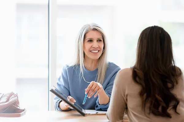 A professional woman sits across a desk from her client, pointing at an iPad and smiling as they have a conversation.