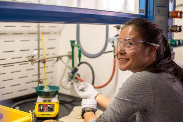 A student works in a campus biology laboratory, amid a variety of hoses and machinery. She wears safety goggles and gloves and holds a syringe.