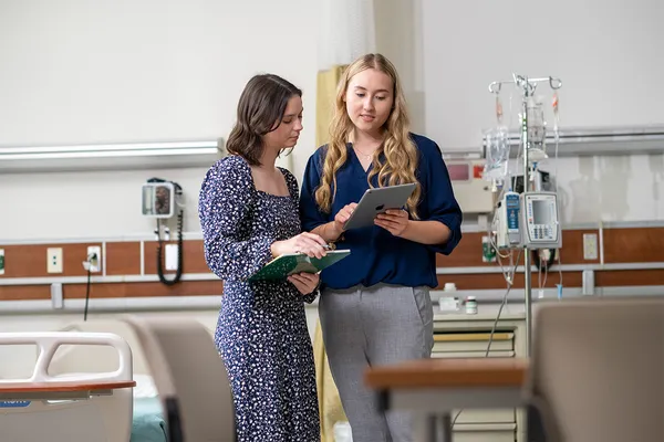 Two young women stand in a medical setting with patient beds and equipment, speaking to one another as one holds a clipboard that they both reference.