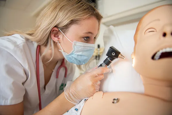 A nursing student wearing a face mask leans over a simulator mannequin, checking its ears with an otoscope.