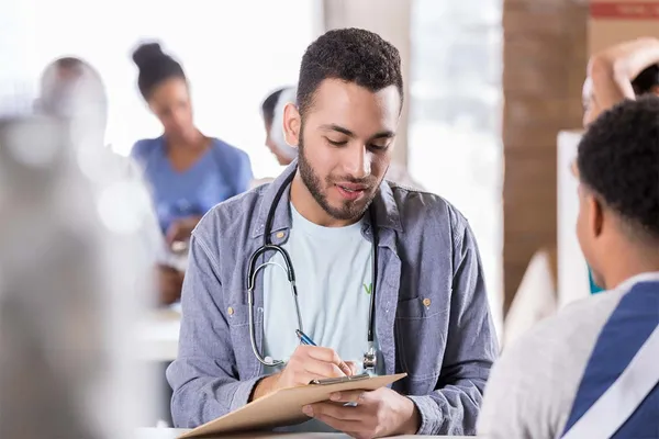 A young man takes notes on a clipboard during a community health event. He wears a stethoscope around his neck.