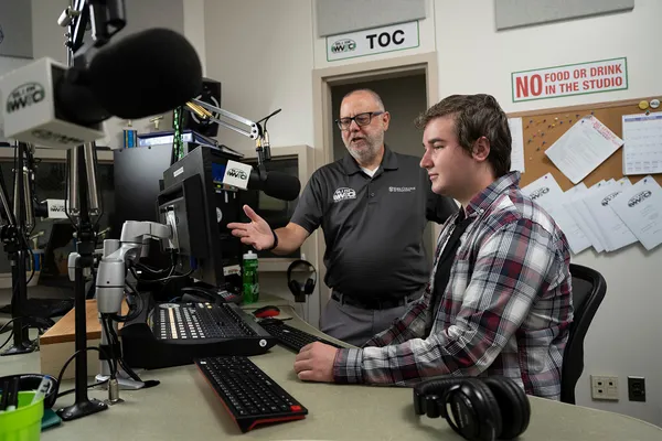Professor Jeff Schiffman discussed equipment in the radio station with a Mass Communication student, surrounded by microphones and other devices.
