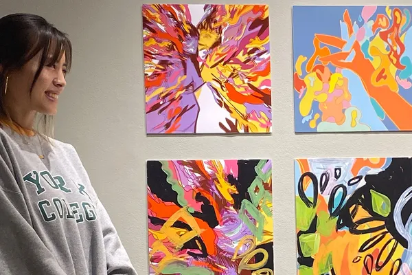 Brittany Dao smiles as she stands beside four colorful paintings on a gallery wall. She wears a York College sweatshirt.