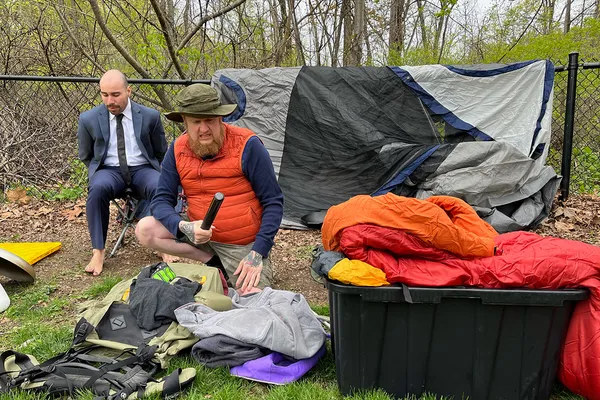 A photo from a scene during an outdoor live play shows two men sitting amid camping gear. One is dressed for hiking; the other wears a suit and is barefoot.