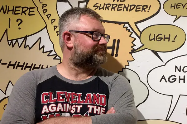 Matthew Borgen stands in front of a wall full of his artwork, a yellow- and gray-toned series of comic book-styles speech bubbles.