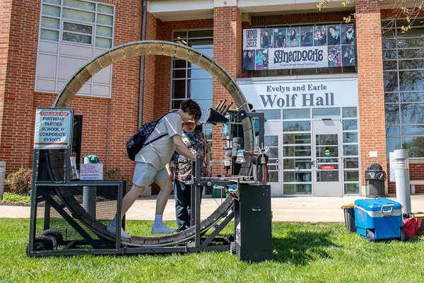 A student walks on a wooden human-sized hamster wheel as the operator looks on on the lawn in front of Wolf Hall. A sign beside the machine reads: Available for birthday parties, school and corporate events.