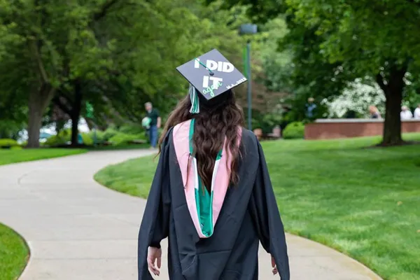A graduating student walks across campus in her regalia, facing away from the camera. Her cap is decorated to read I Did It.