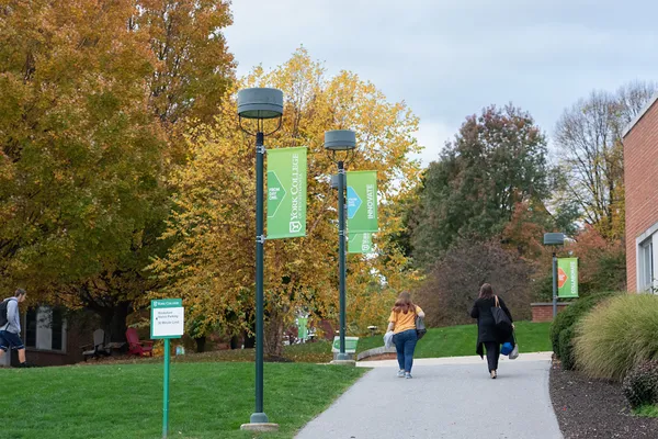 Two students walk away from the camera, following a sidewalk through campus as fall leaves set a colorful scene.