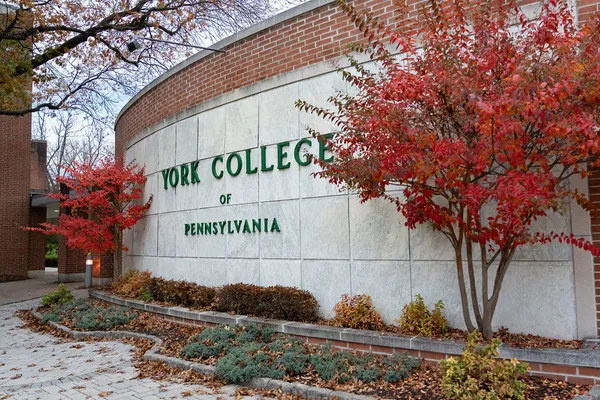 Red fall leaves frame an outdoor stone wall etched with the words York College of Pennsylvania on the campus quad.