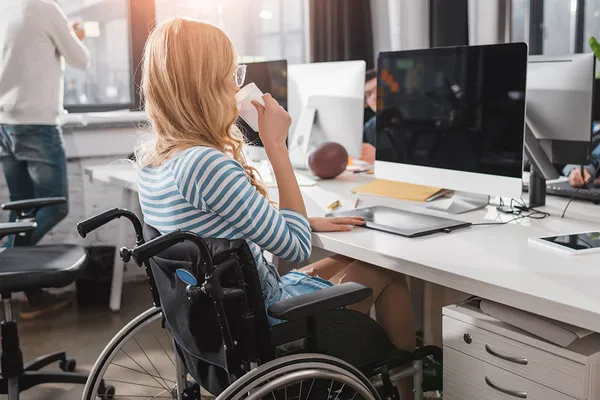 A young woman who is a wheelchair user sits in front of a computer in a computer lab and sips a cup of coffee.