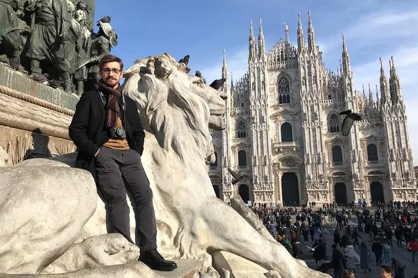 Jack Korver stands in front of a huge lion statue in Italy, with a magnificent gothic architecture cathedral in the background.