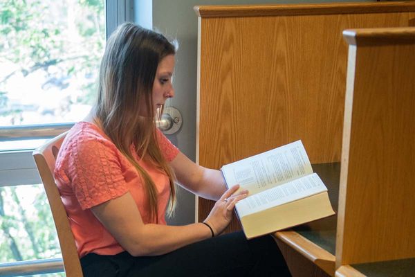 A student sits at a study booth in the library, reading from a large hardcover book.