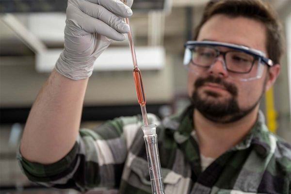 A student holds a pipette full of red liquid while wearing safety goggles and gloves in the lab.