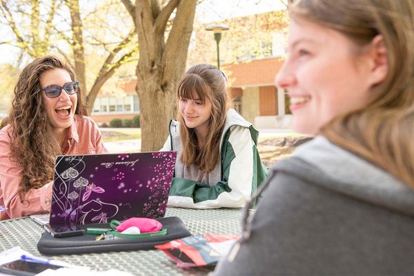 Three students sit outdoors at a picnic table in the campus quad on a brisk spring day. They look at a laptop screen, and each other, and laugh.